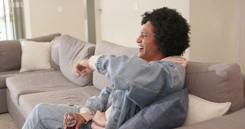African american man laughing and relaxing on gray sofa holding bottle in cozy living room