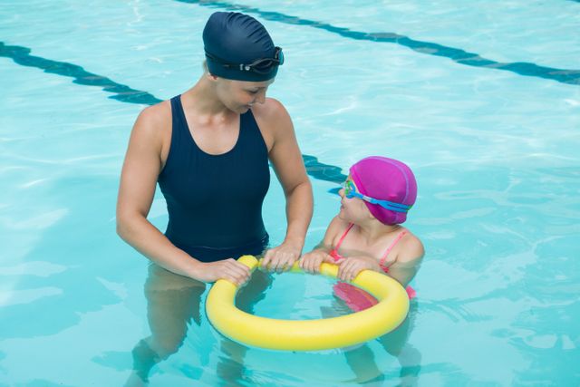 Female instructor training young girl in pool. Both wearing swim caps and goggles. Girl holding flotation device. Ideal for illustrating swim lessons, water safety, child learning to swim, and aquatic activities.