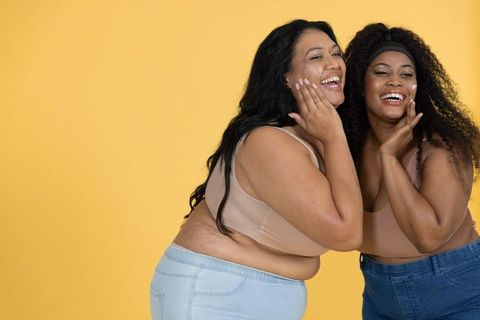 Plus-size friends joyfully laughing together wearing denim and nude tops on yellow background