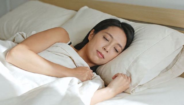 Asian woman resting in bed with neutral bedding, serene intimate morning portrait
