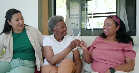 Diverse senior women sharing electric toothbrush, laughing and chatting on sofa