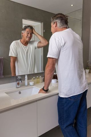 Mature man grooming gray hair at modern bathroom sink looking in mirror