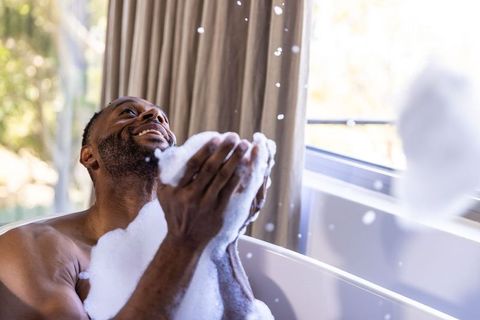 African american man relaxing in sunlit bubble bath holding foam, enjoying calm wellness