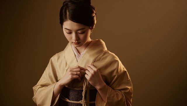 Asian woman adjusting braided cord while wearing beige kimono and dark obi