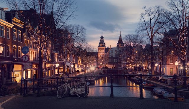 Charming evening scene of Amsterdam with beautifully lit streets and canals. The historic buildings and bicycles parked along the bridge evoke a quintessential European atmosphere. Ideal for use in travel blogs, tourism websites, postcards, and articles highlighting European destinations and nighttime cityscapes.