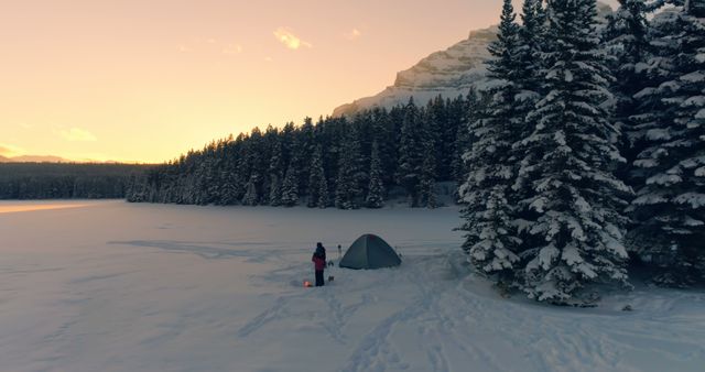 Picture winter camping scene with a person beside a tent in a snow-covered forest at sunset. Ideal for travel blogs, adventure tourism promotions, nature exploration articles, and camping gear advertisements.