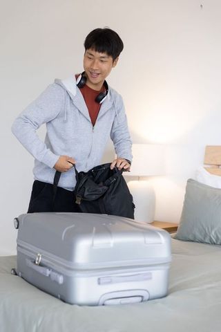 Asian man packing backpack beside silver suitcase in hotel room, preparing for trip