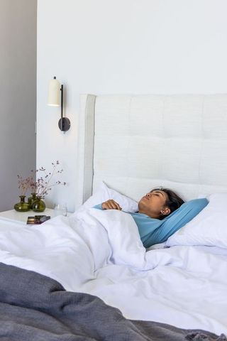 Asian woman resting on bed with tufted headboard, white duvet and gray throw in minimalist bedroom