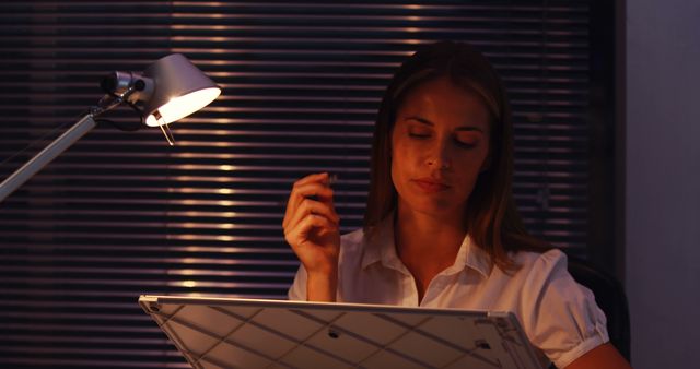 Young professional woman analyzing architectural plans under desk lamp light in an office at night. Ideal for business, corporate late-night, career dedication, and work-life balance visual themes.