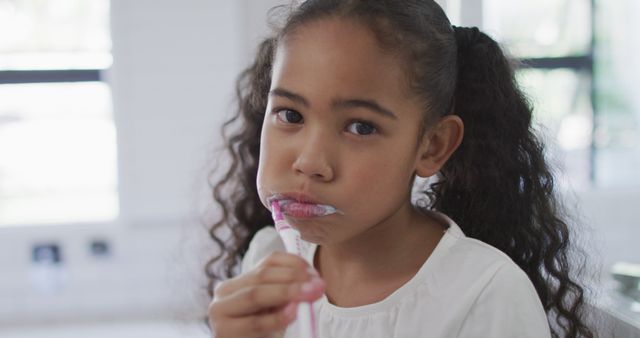 Young girl with curly hair brushing teeth with pink toothbrush in bright bathroom. Promotes good dental hygiene practices, ideal for use in health and wellness articles, parenting blogs, and dental care advertisements.