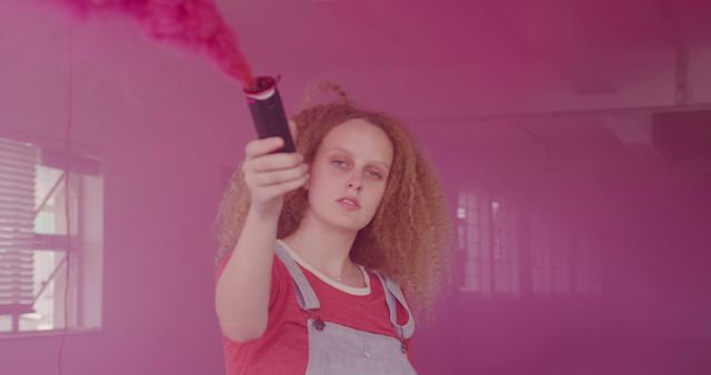 Young woman with curly hair creating vivid pink smoke cloud indoors using smoke bomb. Ideal for creative projects, youth culture promotions, artistic expressions, and urban lifestyle campaigns.