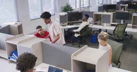 Open-plan office team collaborating across cubicles with monitors and natural daylight