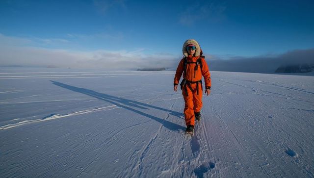 Explorer walking across arctic icefield in orange parka casting long shadow at sunrise