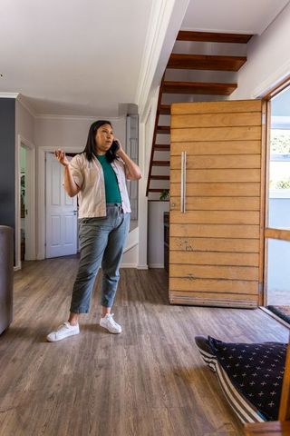 Mature asian woman standing by open wooden front door talking on smartphone in entryway