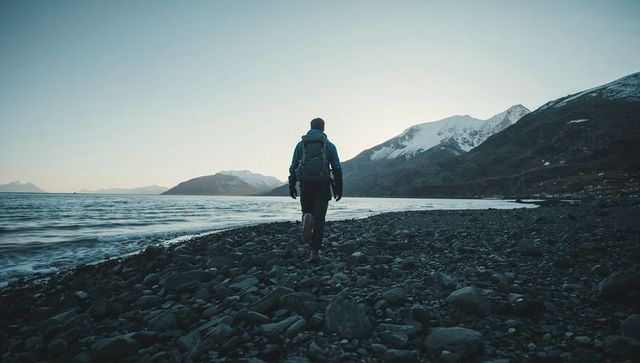 Solo hiker walking along rocky coastline beneath snow-capped mountains at cold dawn
