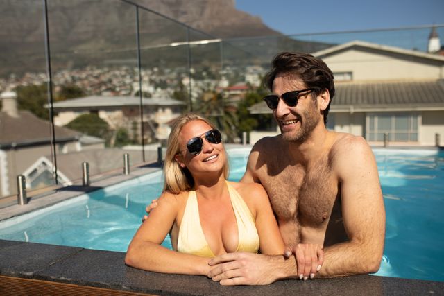 Front view of a happy Caucasian couple enjoying time off in summer at a hotel, wearing sunglasses on a terrace standing in a private swimming pool on a sunny day, embracing and smiling