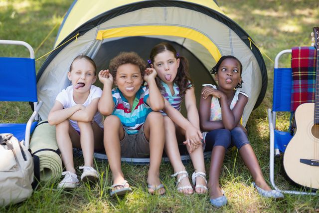 Children sitting in front of a tent making funny faces, enjoying outdoor camping. Perfect for use in advertisements for camping gear, family vacations, outdoor activities, and children's summer camps.