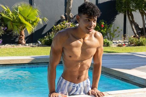 Sunlit african american man emerging from pool in striped trunks, smiling on backyard deck