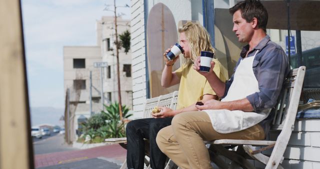 Two friends sitting on a bench outside a coffee shop, drinking hot beverages and engaging in conversation. They appear relaxed and enjoying their time together. The urban setting gives a casual and friendly vibe, ideal for illustrating themes of friendship, relaxation, urban lifestyle, and social interaction.