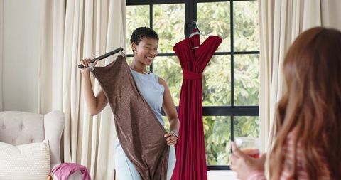 African american woman comparing dresses holding brown dress and smiling by window