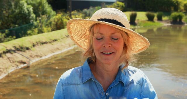 A middle-aged Caucasian woman wearing a straw hat and denim shirt smiles gently in a sunny outdoor setting, with copy space. Her relaxed demeanor suggests a moment of leisure or a break from gardening activities.