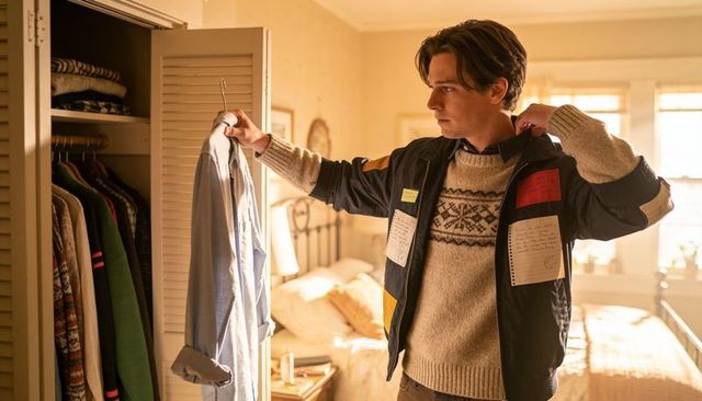 Young man holding shirt while adjusting jacket in sunlit bedroom closet casual wardrobe