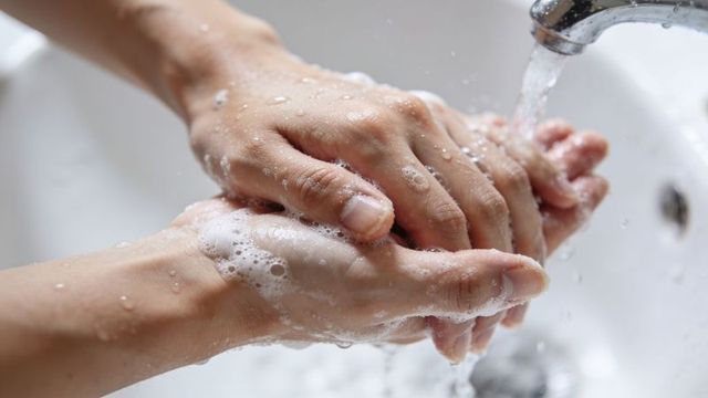 Adult hands washing under running faucet with soap suds and water droplets, closeup