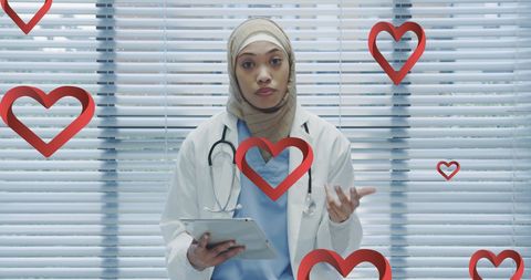Hijabi Doctor Using Tablet with Heart Icons in Hospital