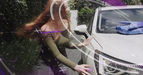 Woman leaning and reaching toward white suv headlight with blue bag on hood in driveway