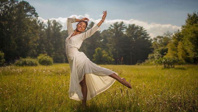 Joyful dancer balancing on one leg in flowing white dress in sunlit meadow