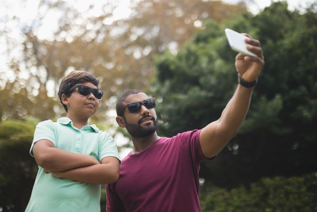 Man and boy standing outdoors, both wearing sunglasses and smiling while taking a selfie with a smartphone. Ideal image for portraying family bonding, outdoor activities, and happy moments. Suitable for articles on parenting, leisure time, and technology in daily life.