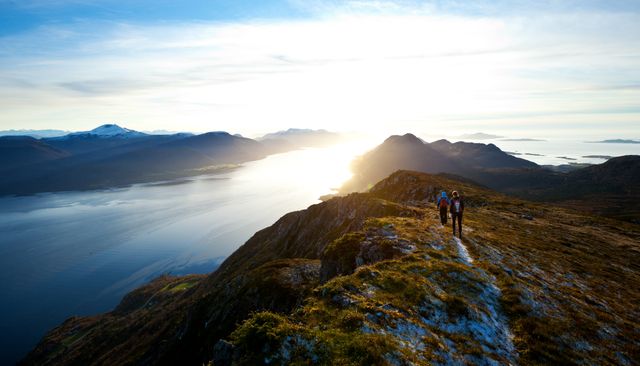 Couple hiking on narrow mountain ridge with sun rising behind distant peaks and calm water body below. Useful for topics on outdoor activities, nature exploration, scenic photography, adventure travel, and fitness. Ideal for travel blogs, adventure catalogs, and promotional materials related to hiking and outdoor excursions.