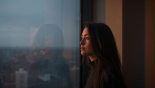 Young woman gazing through city window at sunset, reflective profile in urban apartment