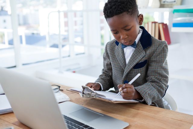 Young boy dressed in a suit working at an office desk, writing in a notebook with a laptop nearby. Ideal for concepts related to business, education, future leaders, and professional development for children.