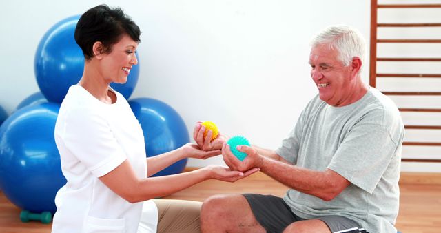 Senior man engaging in a physical therapy session with a smiling therapist. Therapist gently guiding him with therapy balls focusing on motor skill improvement. Useful for illustrating physical therapy practices, senior healthcare services, rehabilitation programs, and healthy aging.