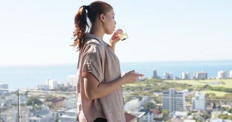Woman drinking on high-rise balcony overlooking coastal cityscape while holding smartphone