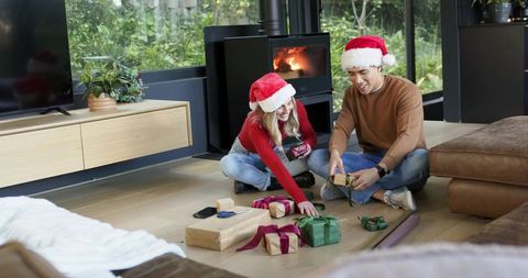 Couple wrapping christmas gifts together wearing santa hats by wood stove fireplace