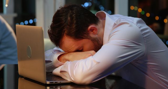 Exhausted businessman sleeping on laptop in office late at night. Looks overworked and fatigued. Suitable for use in articles about work stress, burnout, corporate culture, and the importance of work-life balance.