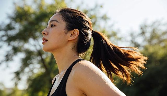 Running asian woman in sunlit park with ponytail and sweat, fitness lifestyle portrait