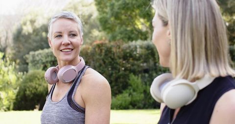 Mature women chatting after workout wearing over-ear headphones in sunny garden wellness moment