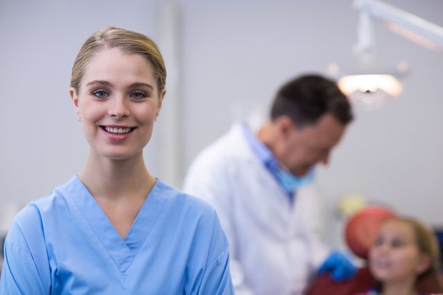 Dental assistant in blue uniform smiling confidently in front of dental equipment and dentist treating young patient in background. Perfect for use in healthcare, dental practice promotions, medical staff training materials, and marketing materials for clinics.