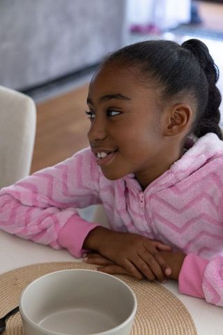African american girl sitting at dining table in pink fleece, smiling and relaxed, cozy morning life
