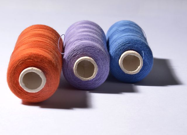 Three colored yarn spools placed on white surface, showing vibrant and contrasting colors of orange, purple, and blue. Ideal for use in sewing, textile, or crafting contexts. Perfect for DIY projects, advertisements for fabric stores, or any creative initiatives involving threads and materials.