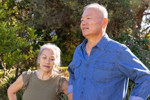 Asian senior couple standing in sunlit garden observing greenery and enjoying quiet moment