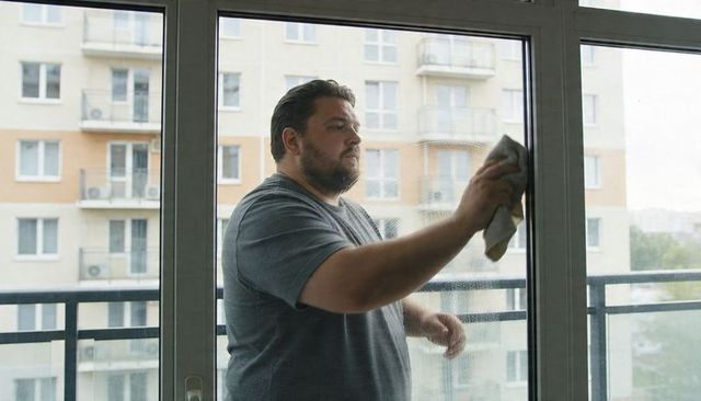 Bearded man cleaning balcony sliding glass door with cloth in urban apartment daytime