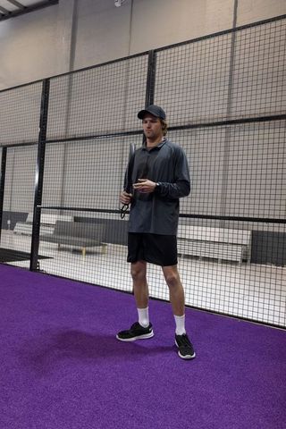Standing athlete holding padel racket on purple indoor court near mesh fence in sportswear