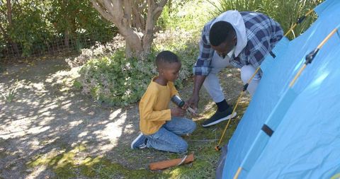 African american father teaching son camping skills pitching blue tent outdoors backyard