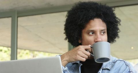 Mid adult african american man sipping from gray mug while working on laptop by window