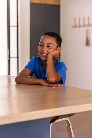 African american boy sitting at wooden table smiling in sunlit modern kitchen