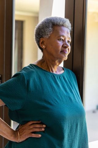 Senior woman standing beside glass door looking thoughtful and serene in sunlit interior